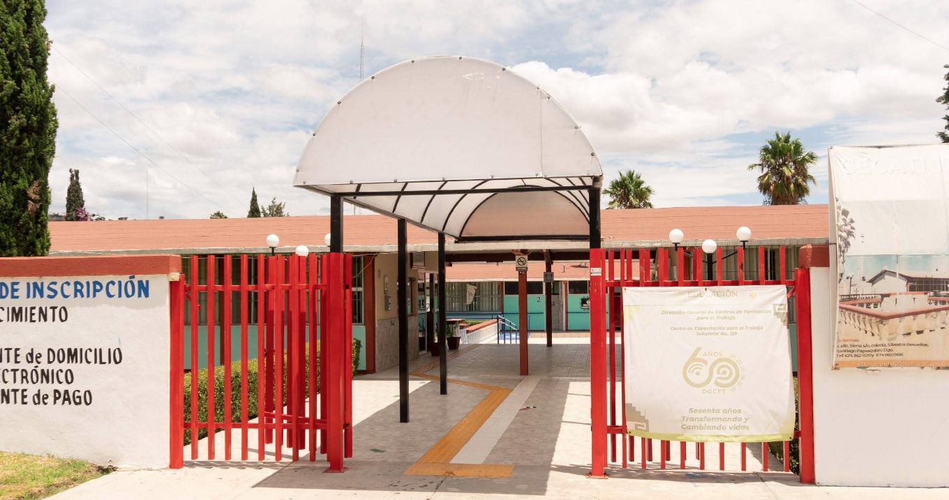 Fotografía en la que se muestra la entrada del plantel, conformada por una reja de color rojo. La reja está abierta, lo que permite observar un pasillo techado. Al terminar el pasillo se encuentran las instalaciones del plantel. 